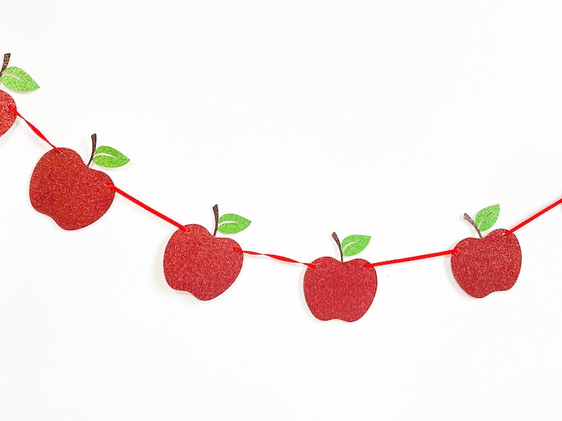 May include: A festive apple garland with red glittery apples strung on a red ribbon. Each apple has a brown stem and a green glittery leaf. The garland is set against a white background.