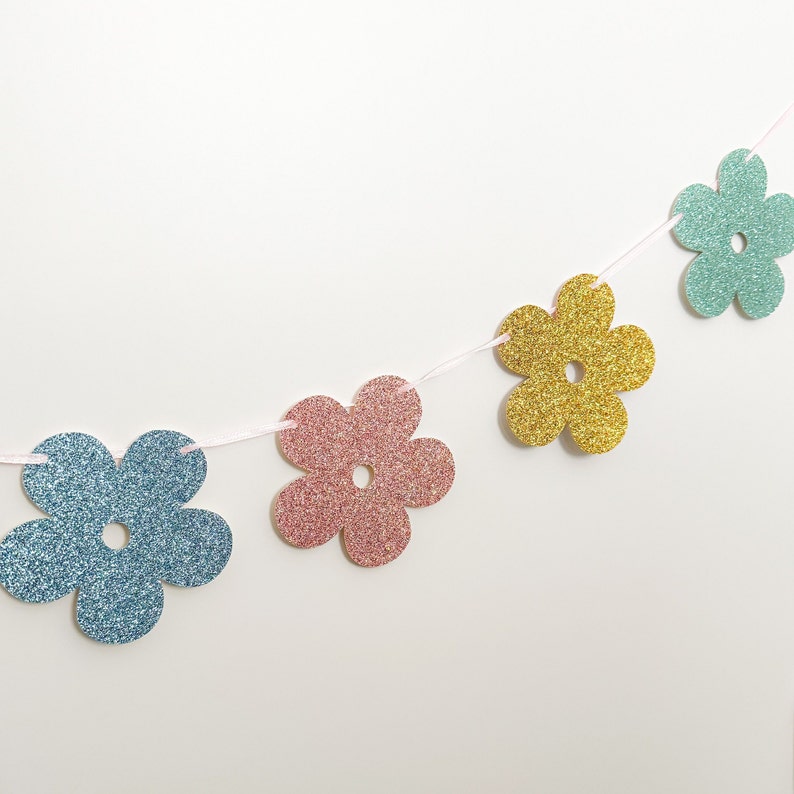 May include: A garland of four glittery flower cutouts in blue, pink, gold, and teal. The flowers are strung on a thin pink ribbon. The flowers have a five-petal design with a small hole in the center of each.