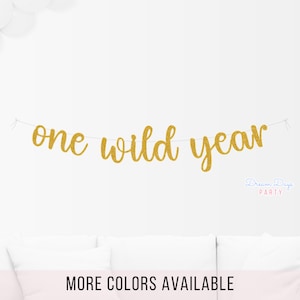 May include: Gold glitter banner with the text "one wild year" hanging on a white wall. The banner is perfect for a first birthday party.