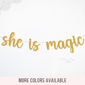 May include: A gold glitter banner that reads "she is magic" strung on a white string. White balloons are in the upper left corner. The words "MORE COLORS AVAILABLE" are at the bottom.