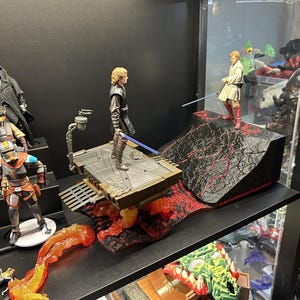 Puede incluir: Una exhibición de figuras de acción de Star Wars. Las figuras de Anakin Skywalker y Obi-Wan Kenobi posan con sables de luz sobre un paisaje volcánico. Otras figuras incluyen a Darth Maul y un Mandaloriano.