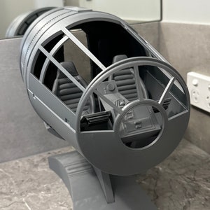 Könnte beinhalten: Ein graues 3D-gedrucktes Modell des Millennium Falcon Cockpits. Das Modell ist detailliert mit einem Bedienfeld, Sitzen und einem Sichtfenster. Das Modell ist auf einem grauen Sockel montiert.