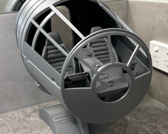 May include: A gray 3D printed model of the Millennium Falcon cockpit. The model is detailed with a control panel, seats, and a viewport. The model is mounted on a gray base.