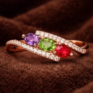 以下が含まれることがあります： 紫、緑、赤の3つの楕円形の宝石が付いたゴールドリング。リングバンドは、小さな透明な石を使った斜めのデザインです。リングは、茶色のテクスチャード加工された背景に配置されています。
