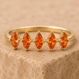 May include: A gold ring featuring five marquise-cut orange gemstones arranged in a horizontal row. The band is smooth and polished, with each vibrant orange stone held securely in place by small metal prongs along the top of the ring setting.