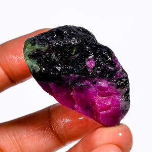 May include: A raw gemstone displaying a dramatic colour gradient. The top section is a deep black, fading into a vibrant magenta at the base. Hints of green are visible near the top, creating a unique mineral specimen.