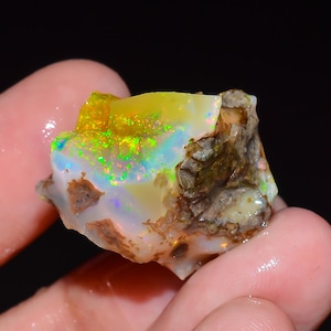 Peut inclure: Une opale brute non polie présentant une base blanc laiteux translucide avec des éclats vibrants de vert néon, de jaune et d'orange. La pierre a une forme irrégulière et dentelée avec des zones de matrice terreuse brune et rugueuse.