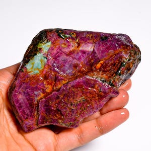 Puede incluir: Una gema de rubí en bruto, sin cortar, de un profundo color magenta, con áreas de naranja, verde y negro. La piedra natural se sostiene en una mano, mostrando su forma irregular y superficie texturizada. Un espécimen mineral único.