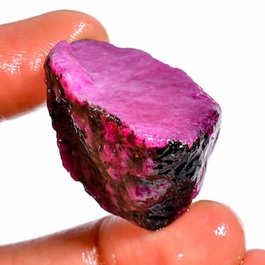 May include: A raw, uncut ruby gemstone, held between fingers. The ruby displays a vibrant pinkish-red hue with dark, almost black, edges. The surface has a rough, natural texture, showcasing the stone's unrefined state. The gemstone is set against a white background.