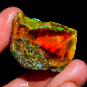May include: A raw, unpolished opal gemstone displaying vibrant flashes of red, orange, and green color play. The stone features a rough, textured surface with embedded mineral inclusions and a translucent, glowing interior against a dark background.