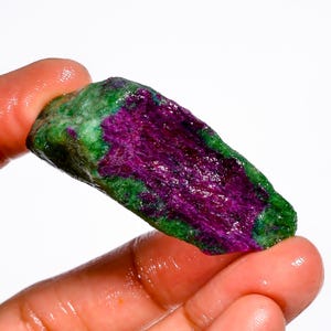 May include: A raw gemstone with a striking colour scheme. The stone displays a deep magenta core, edged with a band of vivid green. The natural texture and colour variations create a striking visual appeal. The gemstone is held in a hand.