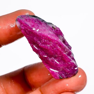 May include: A raw ruby gemstone, displaying a rich, deep pink colour with areas of darker magenta and black. The rough surface of the stone is visible, with a slightly translucent appearance. The gemstone is held against a white background.