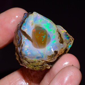 May include: A raw, unpolished opal gemstone featuring a translucent, milky white surface with vibrant flashes of neon green, blue, and orange play-of-color. The center contains a smooth, amber-toned cavity surrounded by rough, earthy rock matrix.