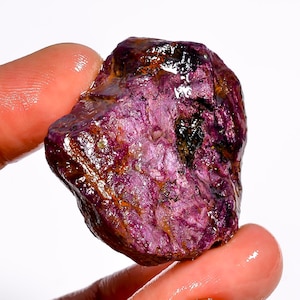 May include: A raw ruby gemstone, displaying a deep magenta hue with a reflective, crystalline surface. The uncut mineral shows a mix of dark red, purple, and brown tones, with visible natural textures and imperfections. The gemstone is held between two fingers.