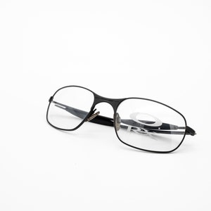 K&ouml;nnte beinhalten: Schwarze Metallgestellbrille mit klaren Gl&auml;sern. Der Markenname "Oakley" ist auf dem rechten Glas sichtbar.