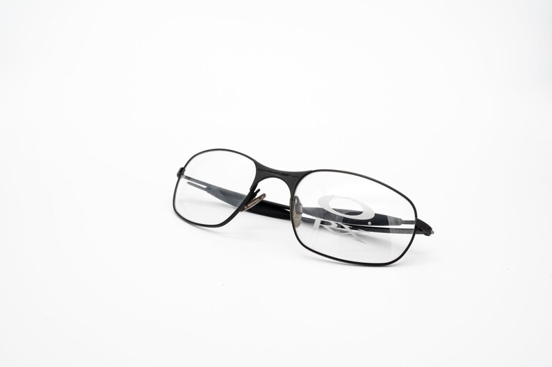 K&ouml;nnte beinhalten: Schwarze Metallgestellbrille mit klaren Gl&auml;sern. Der Markenname "Oakley" ist auf dem rechten Glas sichtbar.