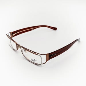 May include: Brown and gold-toned spectacles with a rectangular frame. The arms are a dark brown colour, and the Ray-Ban logo is visible on the arm. The lenses are clear. The glasses are set against a white background.