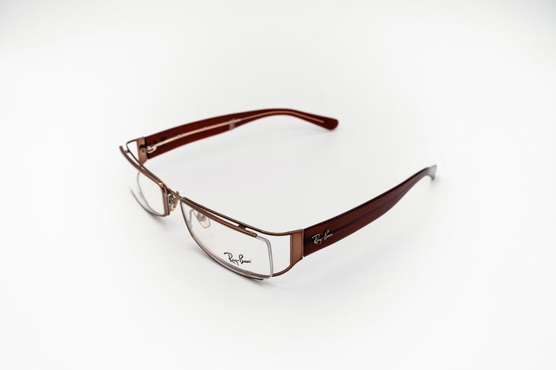 May include: Brown and gold-toned spectacles with a rectangular frame. The arms are a dark brown colour, and the Ray-Ban logo is visible on the arm. The lenses are clear. The glasses are set against a white background.