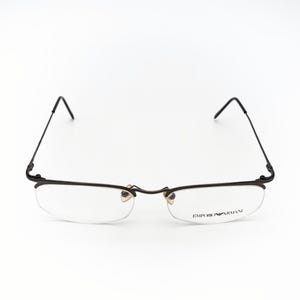 May include: Rimless eyeglasses with a black metal frame. The lenses are clear, and the brand name "EMPORIO ARMANI" is printed on the right lens. The glasses have a minimalist design.
