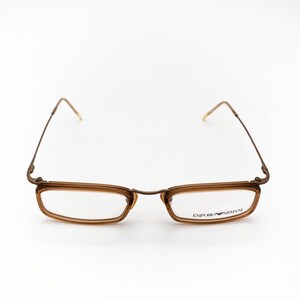 May include: Brown rectangular eyeglasses with thin metal frames and temples. The lenses are clear, and the brand name "EMPORIO ARMANI" is printed on the right lens. The glasses are set against a white background.