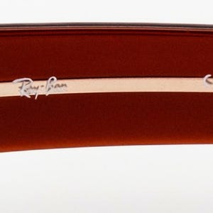 May include: Close-up of the arm of brown sunglasses. The Ray-Ban logo is in silver lettering on a light tan strip. The CE mark is also visible.