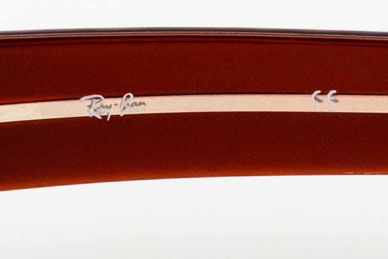 May include: Close-up of the arm of brown sunglasses. The Ray-Ban logo is in silver lettering on a light tan strip. The CE mark is also visible.