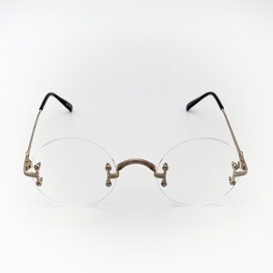 May include: Rimless, round spectacles with a silver-coloured bridge and arms. The lenses are clear, and the earpieces are black. The glasses are set against a white background.