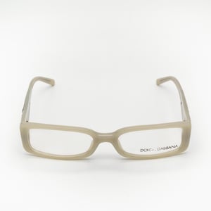 May include: Beige eyeglasses with a rectangular frame and clear lenses. The brand name "Dolce & Gabbana" is printed on the right lens.