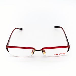 May include: Red and black eyeglasses with a half-rim design. The frames are red with black temples. The lenses are clear, and the right lens has the text "m/m" in red. The glasses are set against a white background.