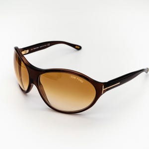 Może przedstawiać: Brązowe okulary przeciwsłoneczne z tworzywa sztucznego z brązowymi szkłami i złotymi akcentami. Okulary mają napis "TOM FORD" nadrukowany na prawym szkle.