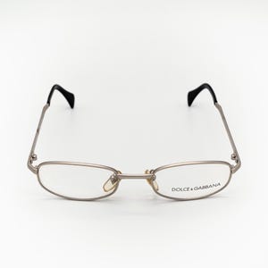Könnte beinhalten: Silberfarbene Metallbrille mit schwarzen Bügeln und einem rechteckigen Gestell. Die Gläser sind klar und der Text "DOLCE & GABBANA" ist auf dem rechten Glas aufgedruckt.