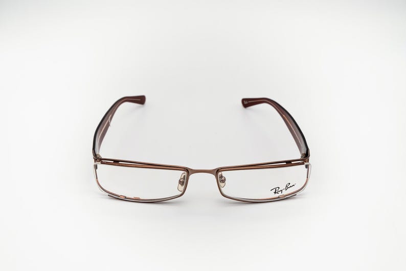 May include: Rectangular spectacles with a brown metal frame and temples. The lenses are clear, and the Ray-Ban logo is visible on the right lens. The spectacles are set against a white background.