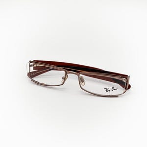 May include: Ray-Ban eyeglasses in brown and gold tones with a rectangular frame. The glasses feature a modern design and the Ray-Ban logo is visible on the right lens. The frames combine brown and gold, offering a sophisticated appearance.