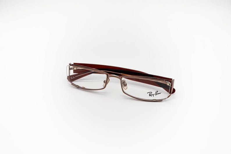 May include: Ray-Ban eyeglasses in brown and gold tones with a rectangular frame. The glasses feature a modern design and the Ray-Ban logo is visible on the right lens. The frames combine brown and gold, offering a sophisticated appearance.
