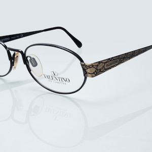 Può includere: Occhiali da vista ovali neri con montatura in metallo dorato. La montatura ha un design testurizzato e le parole "VALENTINO LES LUNETTES" sono stampate sul lato.