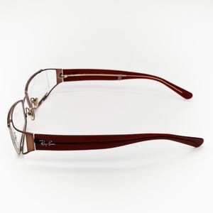 May include: Ray-Ban spectacles with a brown metal frame and dark red arms. The glasses are displayed horizontally against a white backdrop, highlighting their design and the Ray-Ban logo.