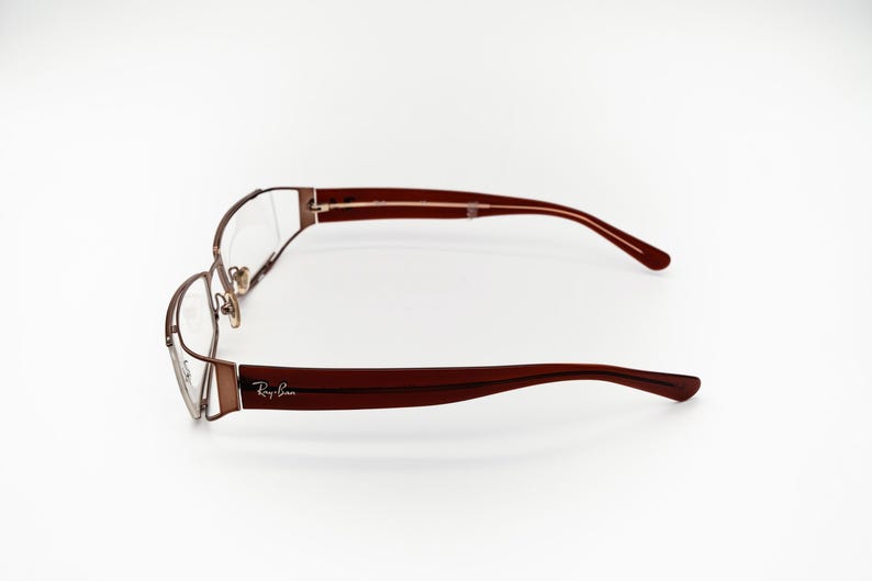 May include: Ray-Ban spectacles with a brown metal frame and dark red arms. The glasses are displayed horizontally against a white backdrop, highlighting their design and the Ray-Ban logo.