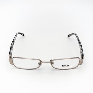 Op de afbeelding: Zilverkleurige DKNY-bril met rechthoekige glazen. De monturen hebben een modern design, met zwart-wit patroon op de pootjes. Het DKNY-logo is zichtbaar op het rechterglas.