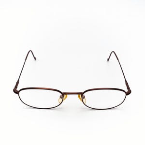 以下が含まれることがあります： 細いダークブラウンの金属フレームの楕円形の眼鏡。レンズは透明で、鼻ブリッジは明るい金色です。テンプルもダークブラウンで、端がわずかに湾曲しています。