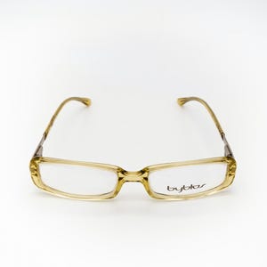 May include: A pair of eyeglasses with a light brown frame and clear lenses. The rectangular frames have a subtle, translucent quality. The brand name "byblos" is printed on the right lens.
