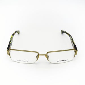 May include: Eyeglasses with a gold-colored metal frame and patterned temples. The lenses are clear, and the text "STAINLESS STEEL" and "EMPORIO ARMANI" are visible on the lenses. The glasses are set against a white background.