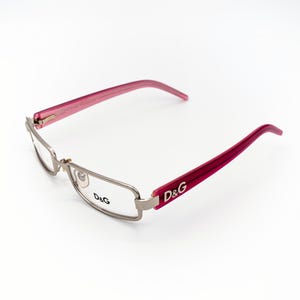 Könnte beinhalten: Brille mit silbernem Rahmen und rosa Bügeln. Das D&G-Logo ist auf dem Bügel sichtbar. Die Brille hat eine rechteckige Linsenform.