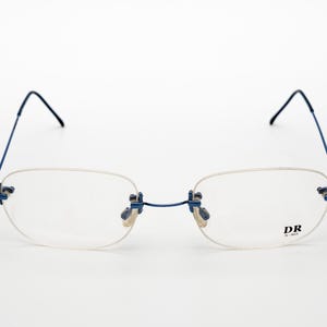 Può includere: Occhiali da vista blu senza montatura con ponte argentato e lenti trasparenti. Il nome del marchio "DR" è stampato sulla stanghetta destra.