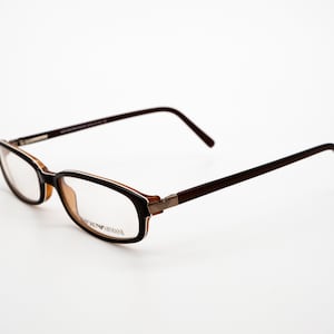 May include: Brown and tan eyeglasses with a silver accent on the temple. The brand name "GIORGIO ARMANI" is printed on the frame.