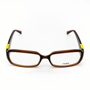 Può includere: Occhiali rettangolari marroni con un accento giallo sulle aste. Gli occhiali hanno una montatura marrone scuro e il marchio "FENDI" è stampato sulla lente destra.