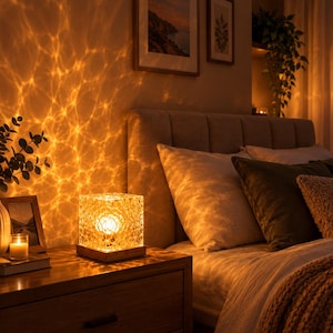 Puede incluir: Una lámpara de mesa cuadrada con un exterior transparente de facetas tipo cristal y una base de madera. La lámpara emite una luz cálida y brillante que proyecta un patrón ondulado similar al agua sobre la pared y las superficies.