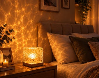 Puede incluir: Una lámpara de mesa cuadrada con un exterior transparente de facetas tipo cristal y una base de madera. La lámpara emite una luz cálida y brillante que proyecta un patrón ondulado similar al agua sobre la pared y las superficies.