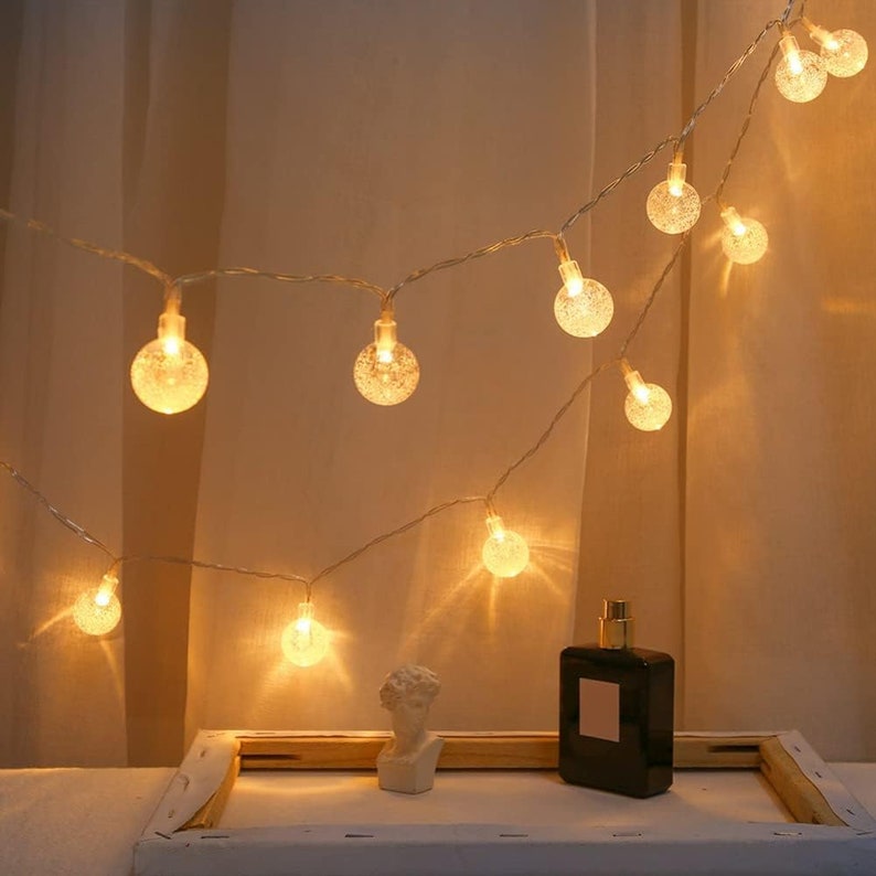Puede incluir: Luces de cadena con peque&ntilde;as bombillas redondas y brillantes. Las luces est&aacute;n colgadas sobre un fondo neutro. Un peque&ntilde;o busto blanco y un frasco de perfume negro est&aacute;n sobre una superficie blanca debajo.