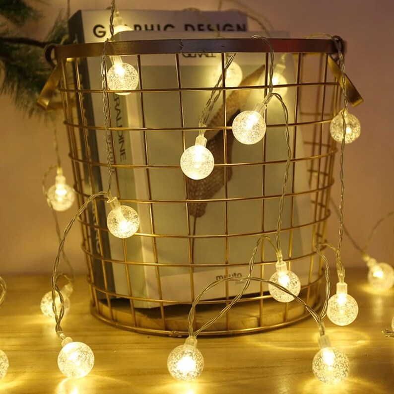 Puede incluir: Una cadena de luces LED de globo blanco c&aacute;lido frente a una cesta de alambre dorada. Las luces son peque&ntilde;as, redondas y emiten un brillo suave. La cesta contiene libros y est&aacute; colocada sobre una superficie de madera, creando un ambiente acogedor.