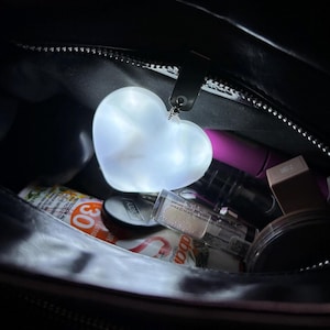 May include: A glowing white heart-shaped keyring hangs inside a black bag. The bag contains various cosmetic items, including sunscreen, lipsticks, and makeup containers. The bag's zipper is visible.